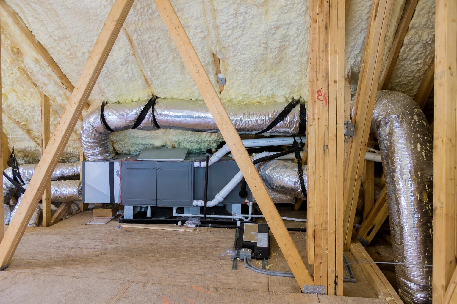 An attic HVAC unit and silver ducts sit beneath roof rafters coated with spray foam insulation.
