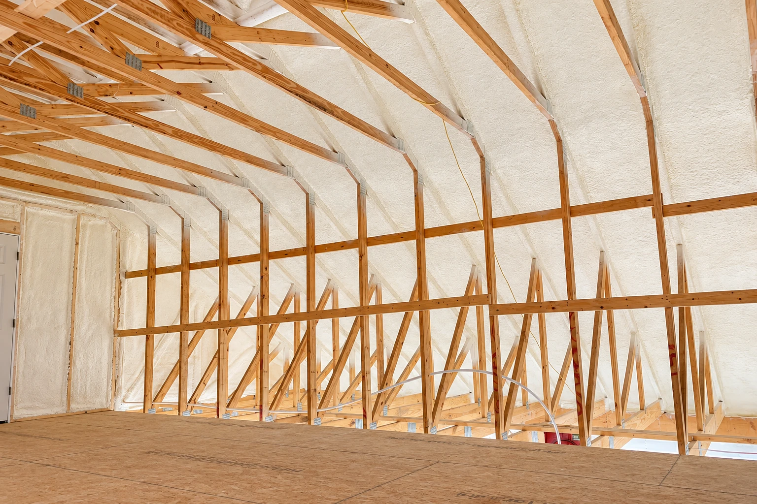 Attic rafters and trusses coated with thick white spray foam above an unfinished plywood floor.