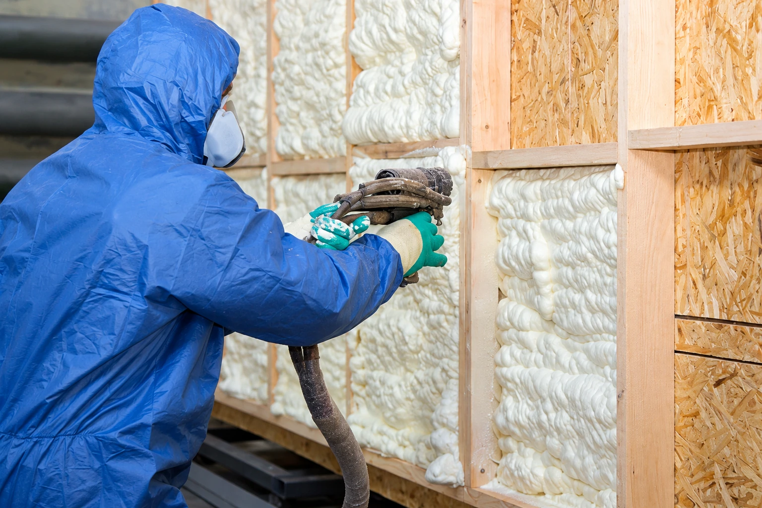 A worker in blue protective gear sprays foam insulation into exposed wall cavities.