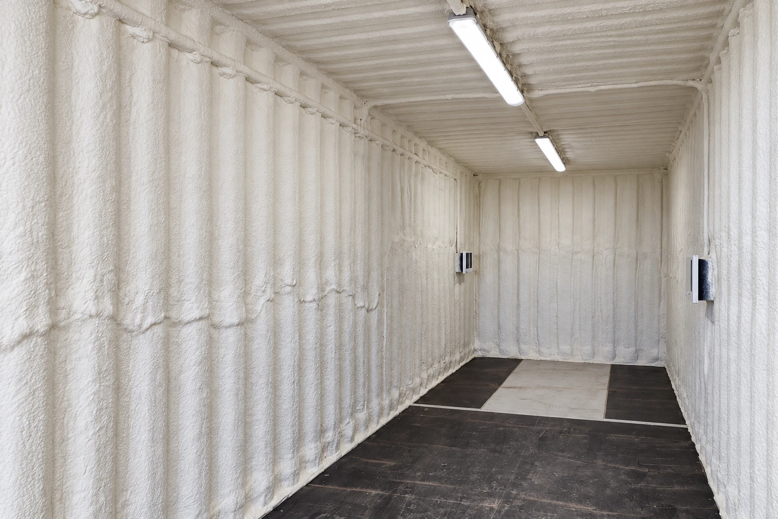 White spray foam coats the walls and ceiling of a narrow commercial room with overhead lights.