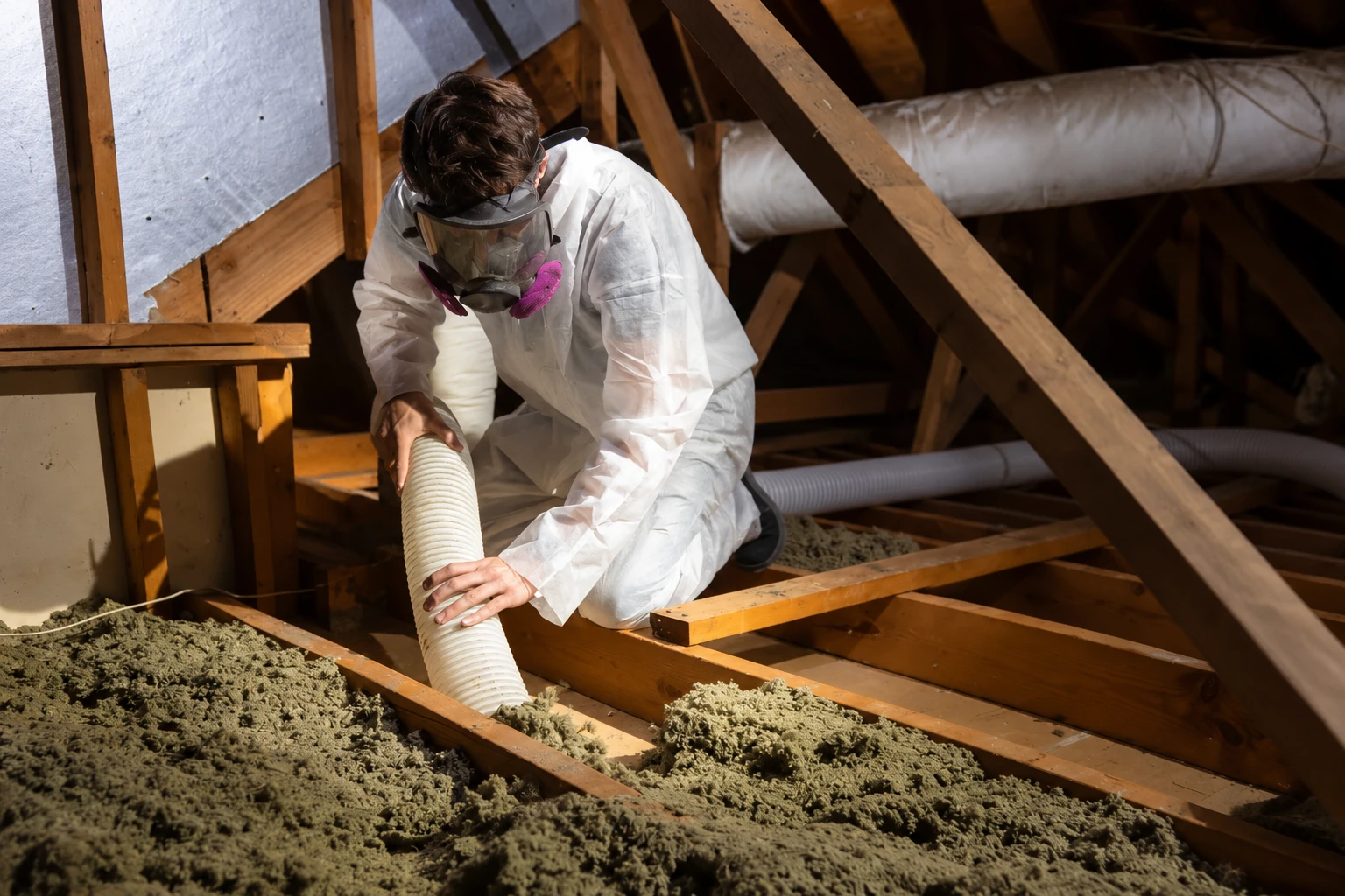 A worker in protective clothing and a respirator vacuums loose attic insulation through a flexible hose.