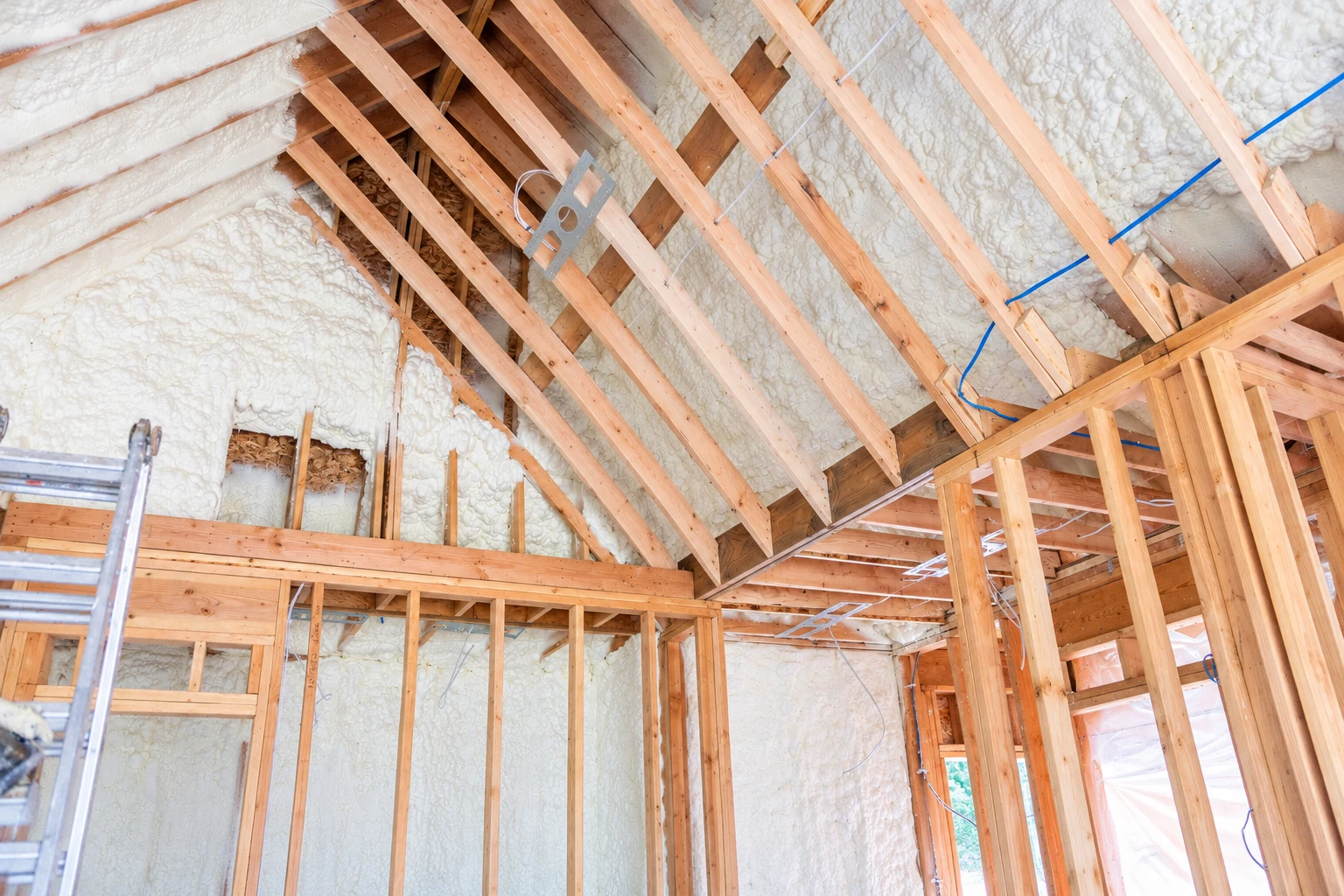 Spray foam coats the roof and wall cavities of an unfinished timber-framed house, with wiring and a ladder visible.